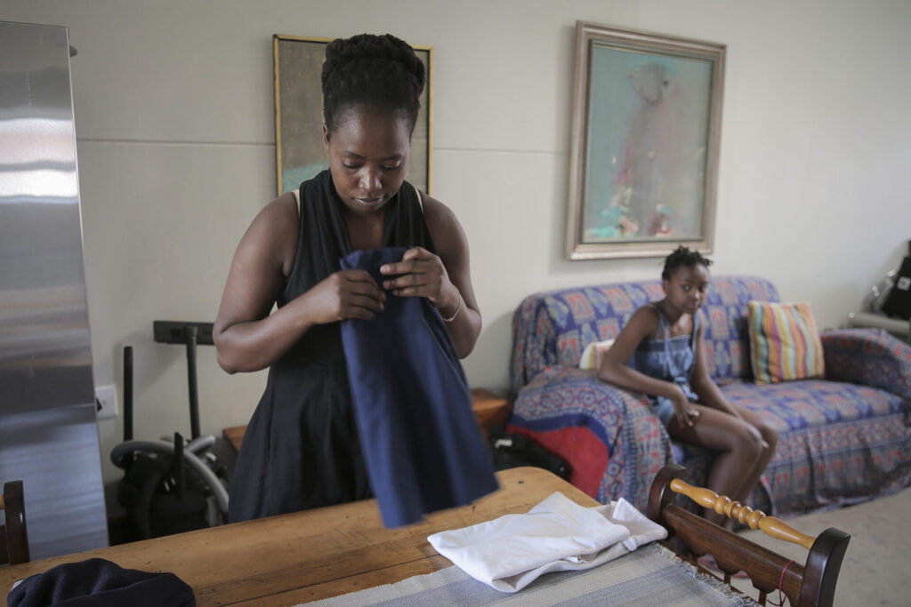 Primrose Modisane folding clothes on a dining table with her eldest daughter Nosipho in the backgroung sitting on a two-seater couch.