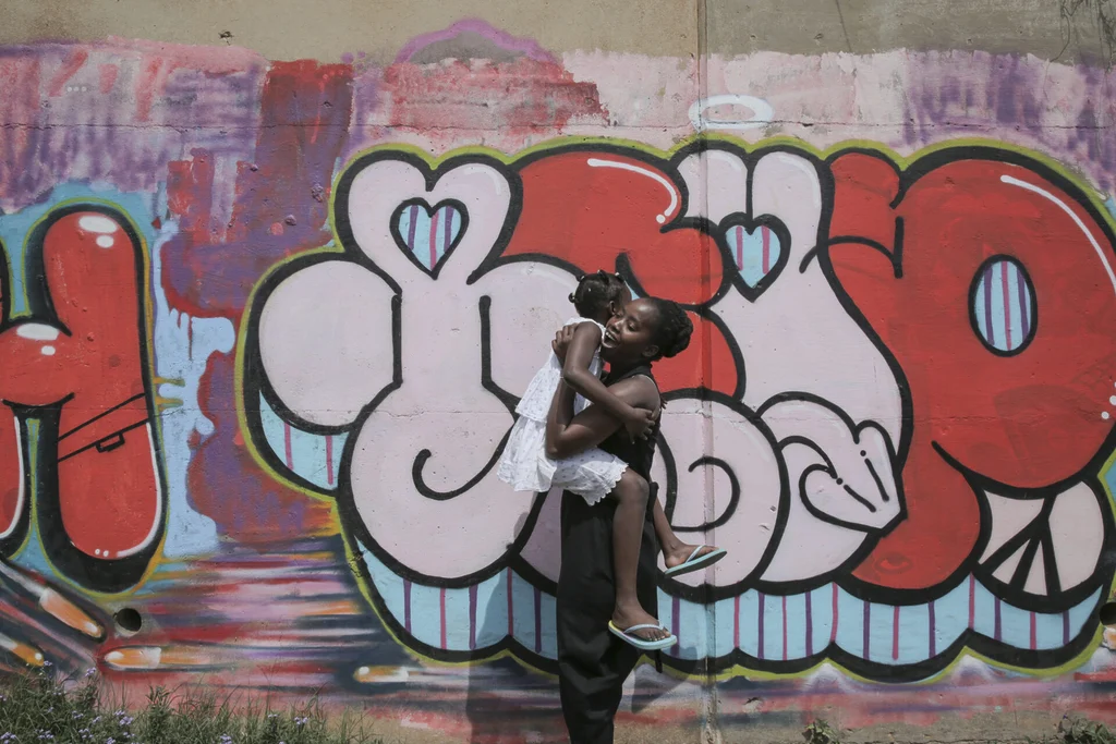 Primrose Modisane holds her child in front of a wall with bright coloured graffiti.